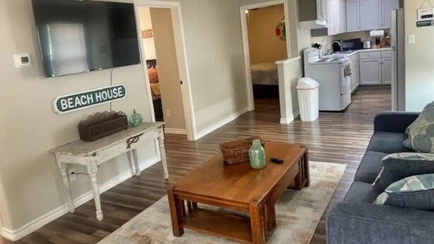 A living room with a wooden coffee table, a sofa, and a flat-screen TV mounted on the wall. There is a sign that reads "BEACH HOUSE" on the wall.