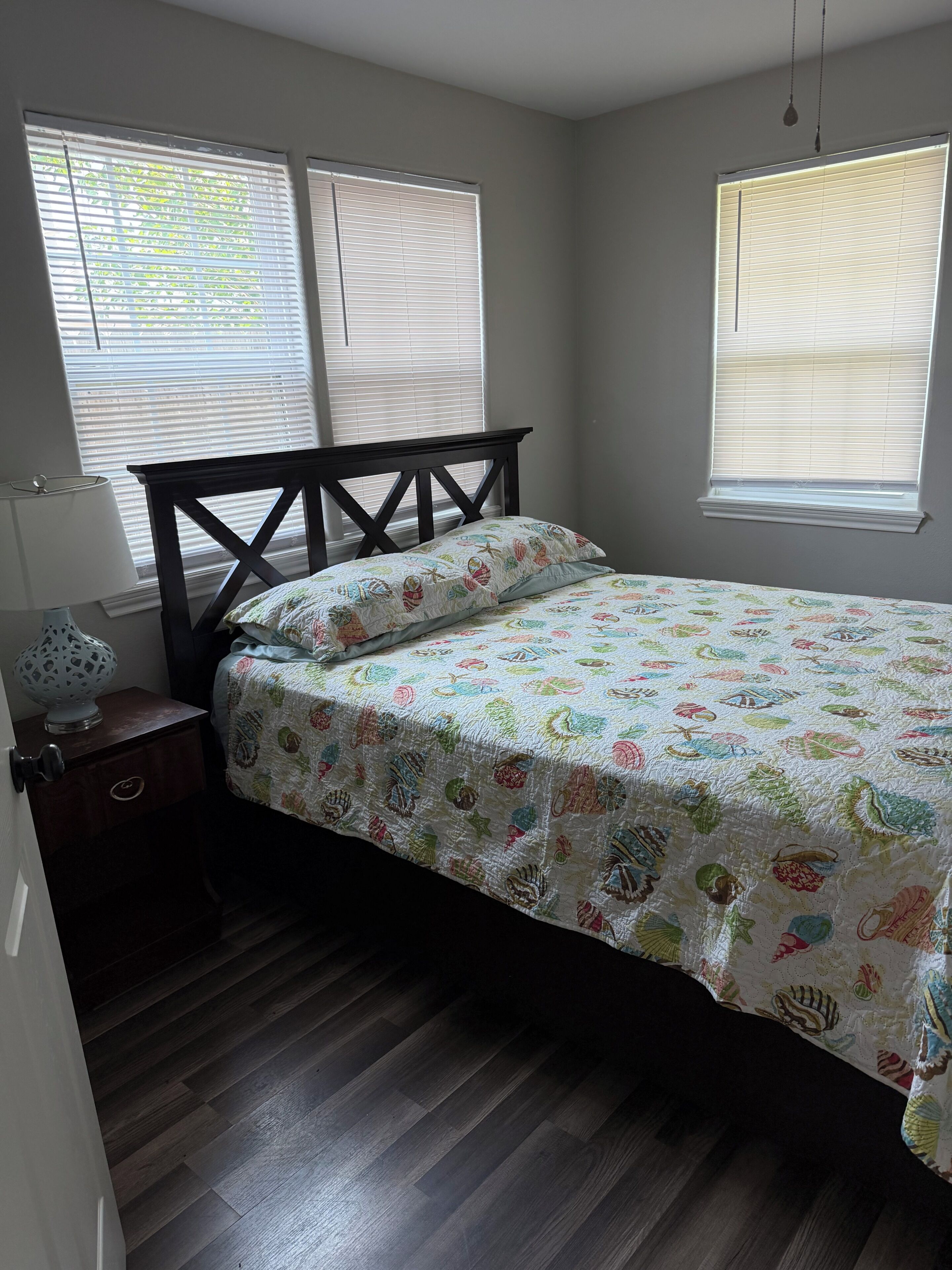 Une chambre à coucher avec un lit, une table de chevet, une lampe et deux fenêtres équipées de stores.