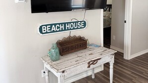 A room with a mounted flat-screen TV, a small table with a decorative basket, and a sign reading 'BEACH HOUSE' on the wall.