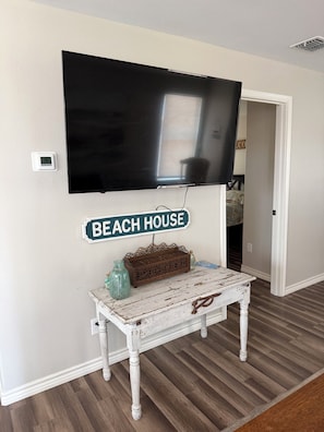 Quarto com TV de tela plana montada, uma mesinha com um cesto decorativo e uma placa na parede com a inscrição 'BEACH HOUSE'.