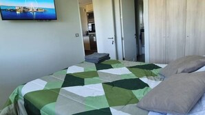 Une chambre à coucher avec un lit, des oreillers, un téléviseur fixé au mur, et une vue sur la cuisine et la salle de bain grâce à une porte ouverte.