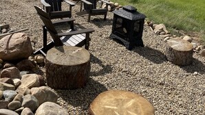 A rustic outdoor seating area with Adirondack chairs and a wood-burning stove, surrounded by a gravel path and large rocks.