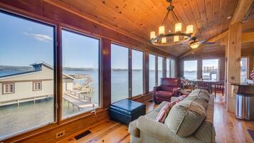 A spacious room with large windows offering a view of a dock and water, wooden flooring, a sofa, and a ceiling fan.