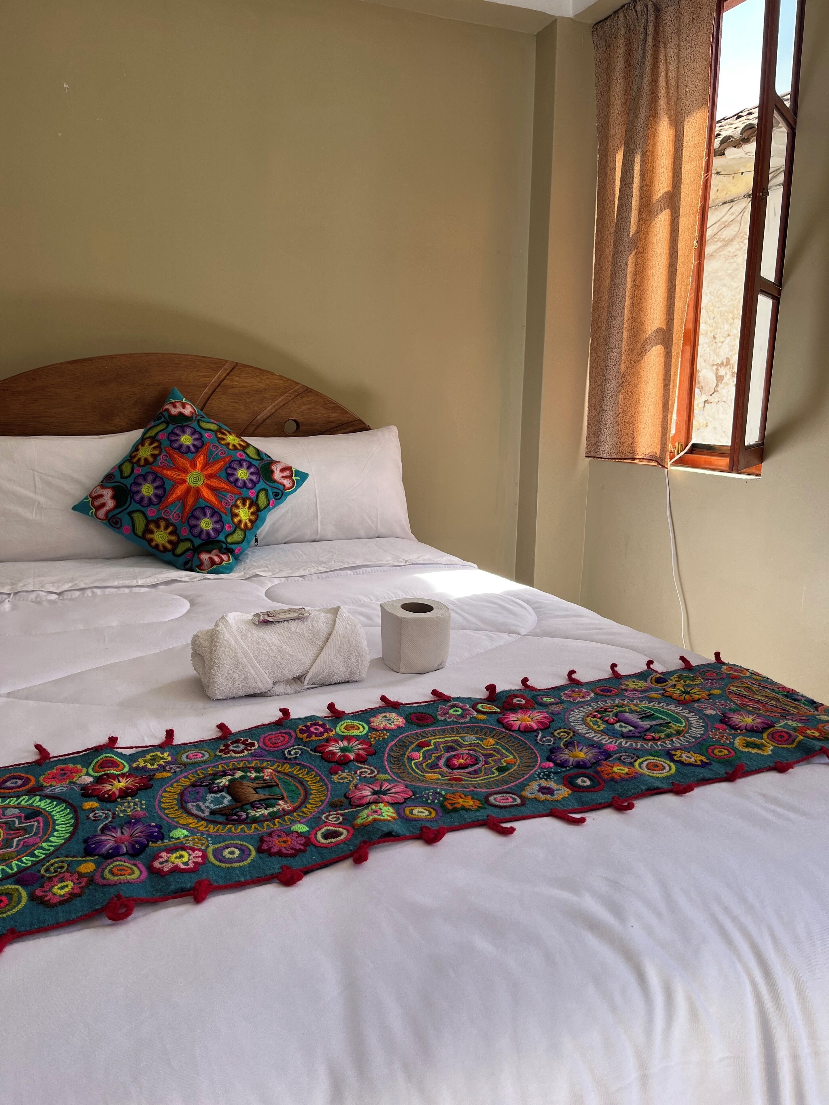 A neatly made bed with a colorful embroidered pillow, a rolled towel, and a decorative rug.