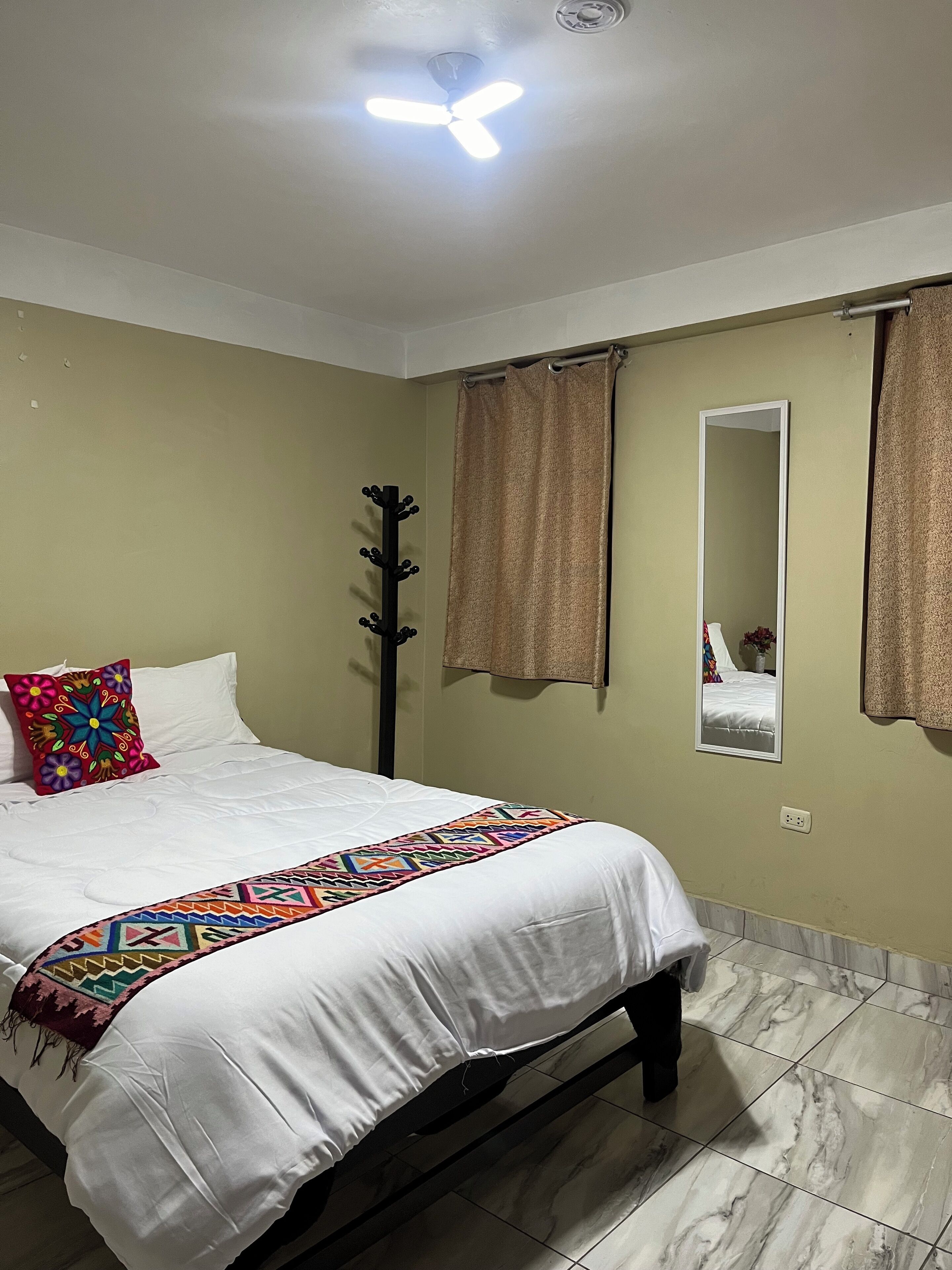 A bedroom with a bed, a ceiling fan, a mirror, and curtains.