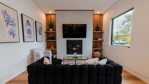 A modern living room with a black sectional sofa, a flat-screen TV, a fireplace, and framed botanical prints on the wall. - Spacious Modern Escape Near UO Campus (Eugene)