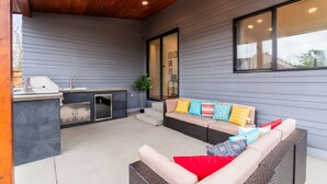 An outdoor patio area with a built-in grill, a seating area with cushions, and a large window. - Spacious Modern Escape Near UO Campus (Eugene)