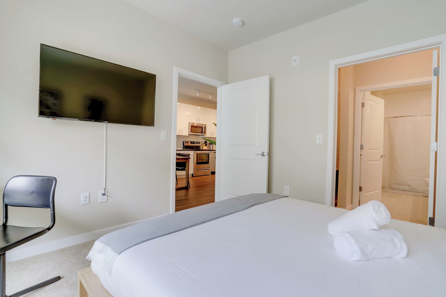 A hotel room with a bed, a TV mounted on the wall, a chair, and an open door leading to a kitchen area.