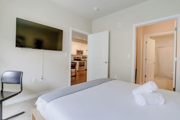A hotel room with a bed, a TV mounted on the wall, a chair, and an open door leading to a kitchen area. - Huntington Apartments near Cameron Run - We Host (Alexandria)