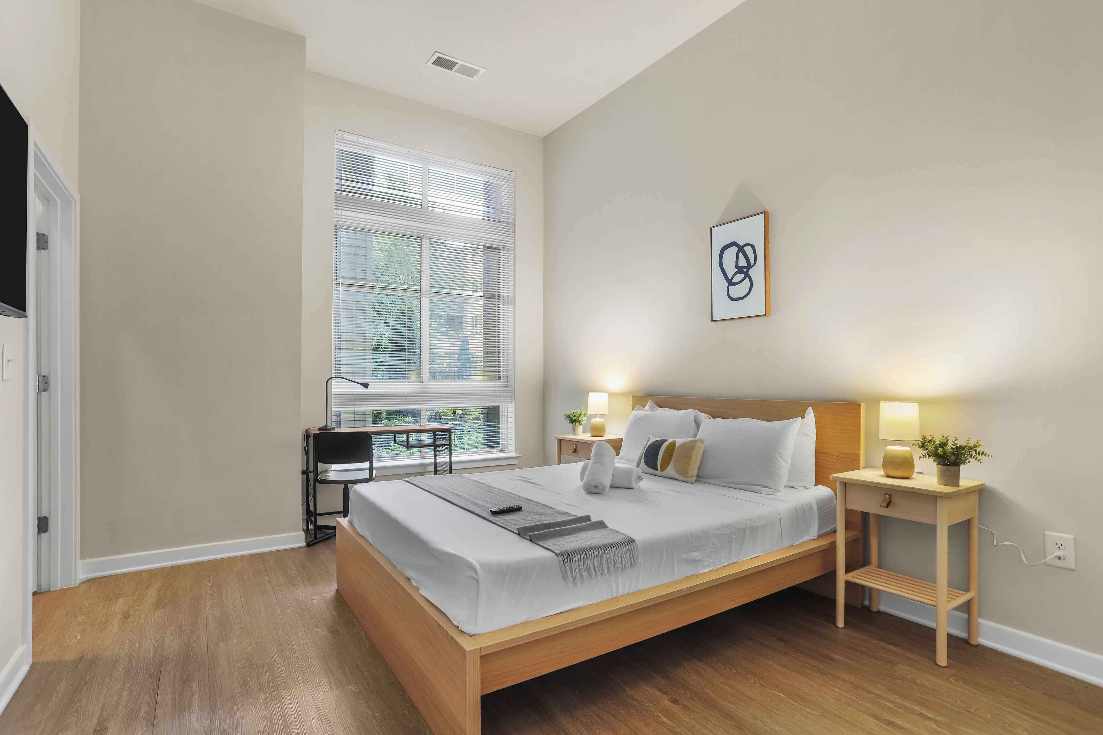 A modern bedroom with a wooden bed, bedside tables, a desk, and a window with blinds.