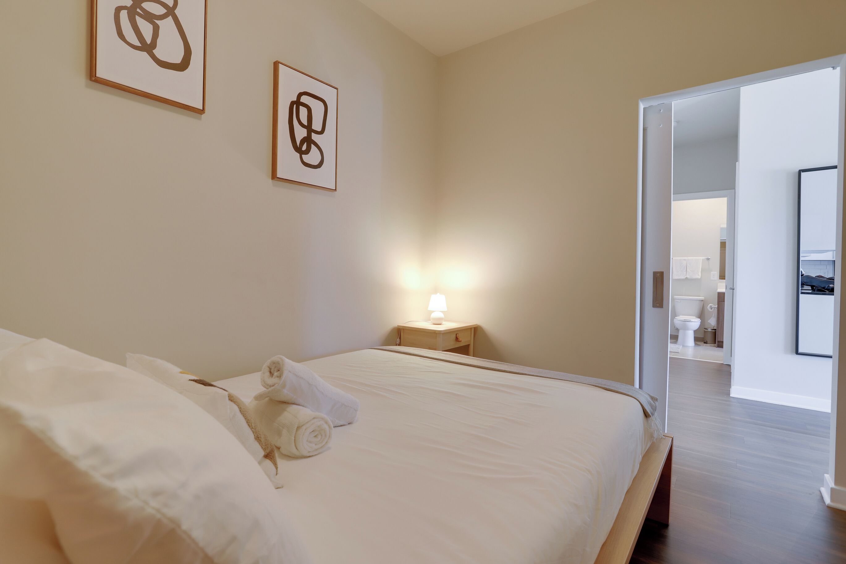 Een net opgemaakt bed met witte lakens en een opgerolde handdoek, een bedlamp en abstracte wanddecoratie.