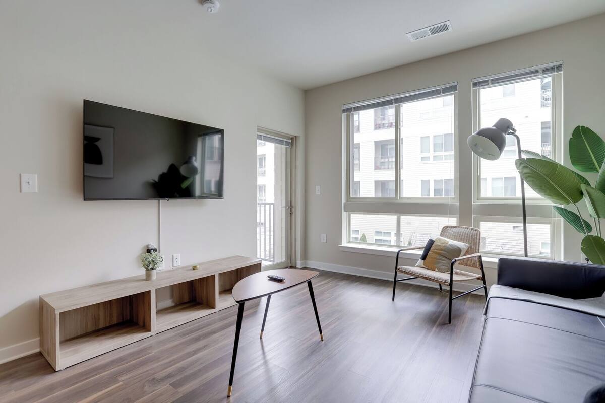 A modern living room with a flat-screen TV, wooden shelves, a coffee table, a sofa, and large windows offering a view of the city.