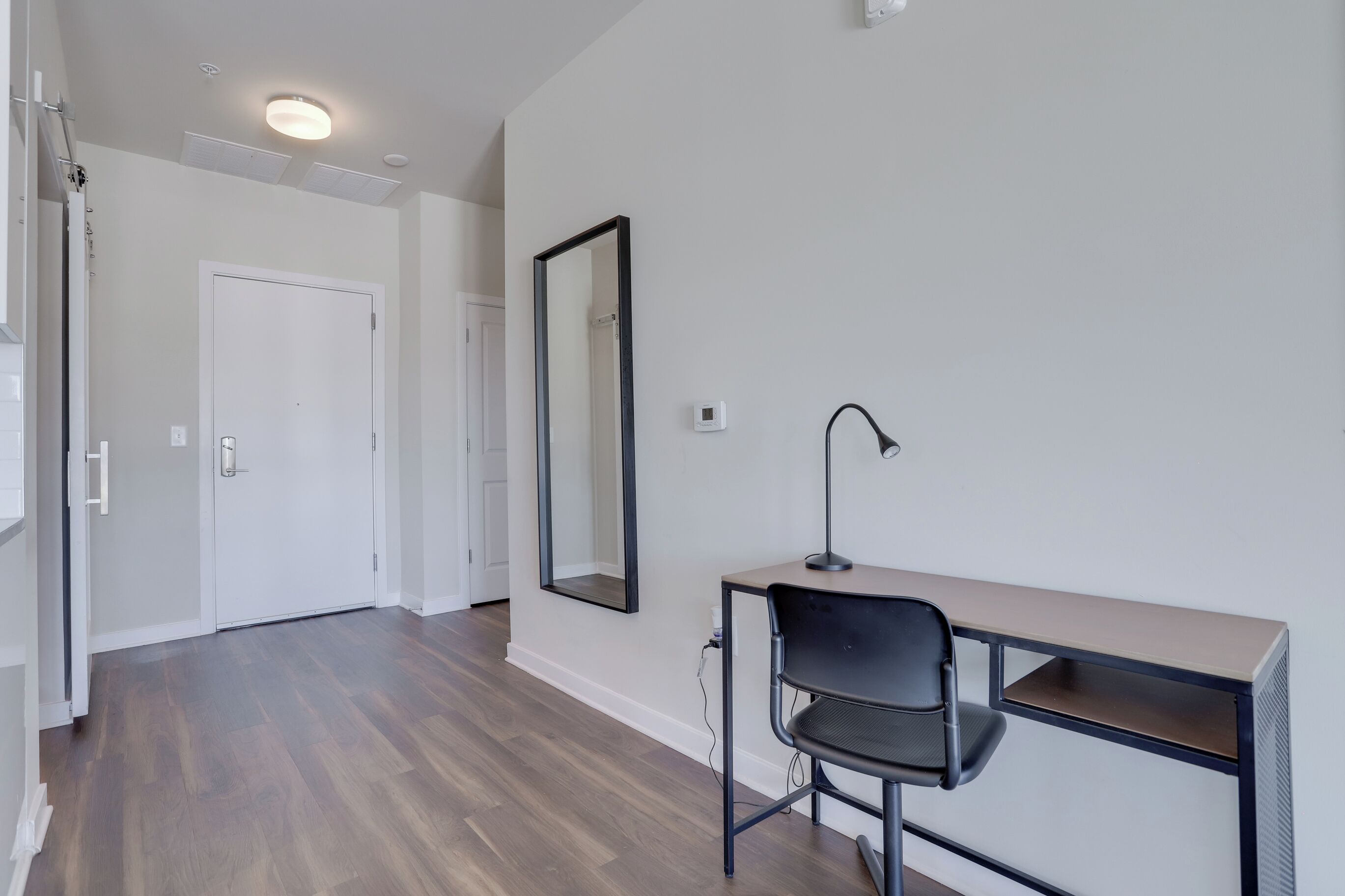 A modern, minimalistic room with a wooden floor, a desk with a chair, a large mirror, and a white door.
