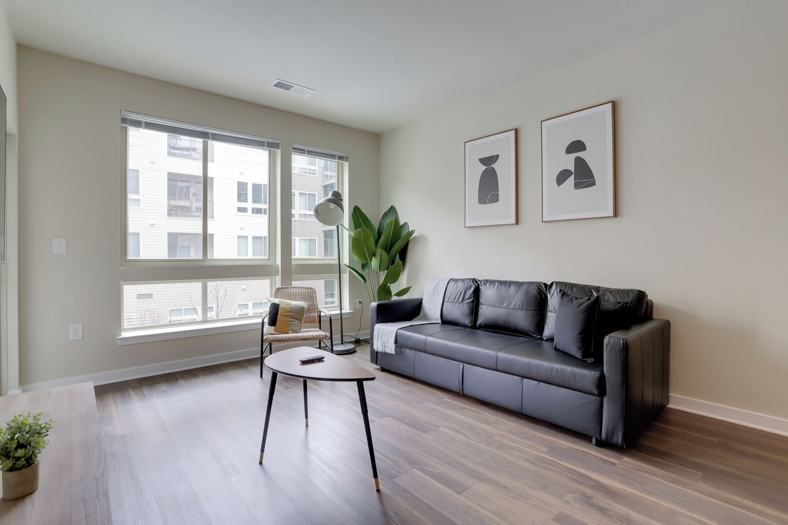 Een moderne woonkamer met een grijze leren bank, een ronde salontafel en een groot raam met zonwering. Twee abstracte kunstwerken hangen in de lijst aan de muur.
