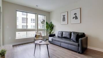 Een moderne woonkamer met een grijze leren bank, een ronde salontafel en een groot raam met zonwering. Twee abstracte kunstwerken hangen in de lijst aan de muur.