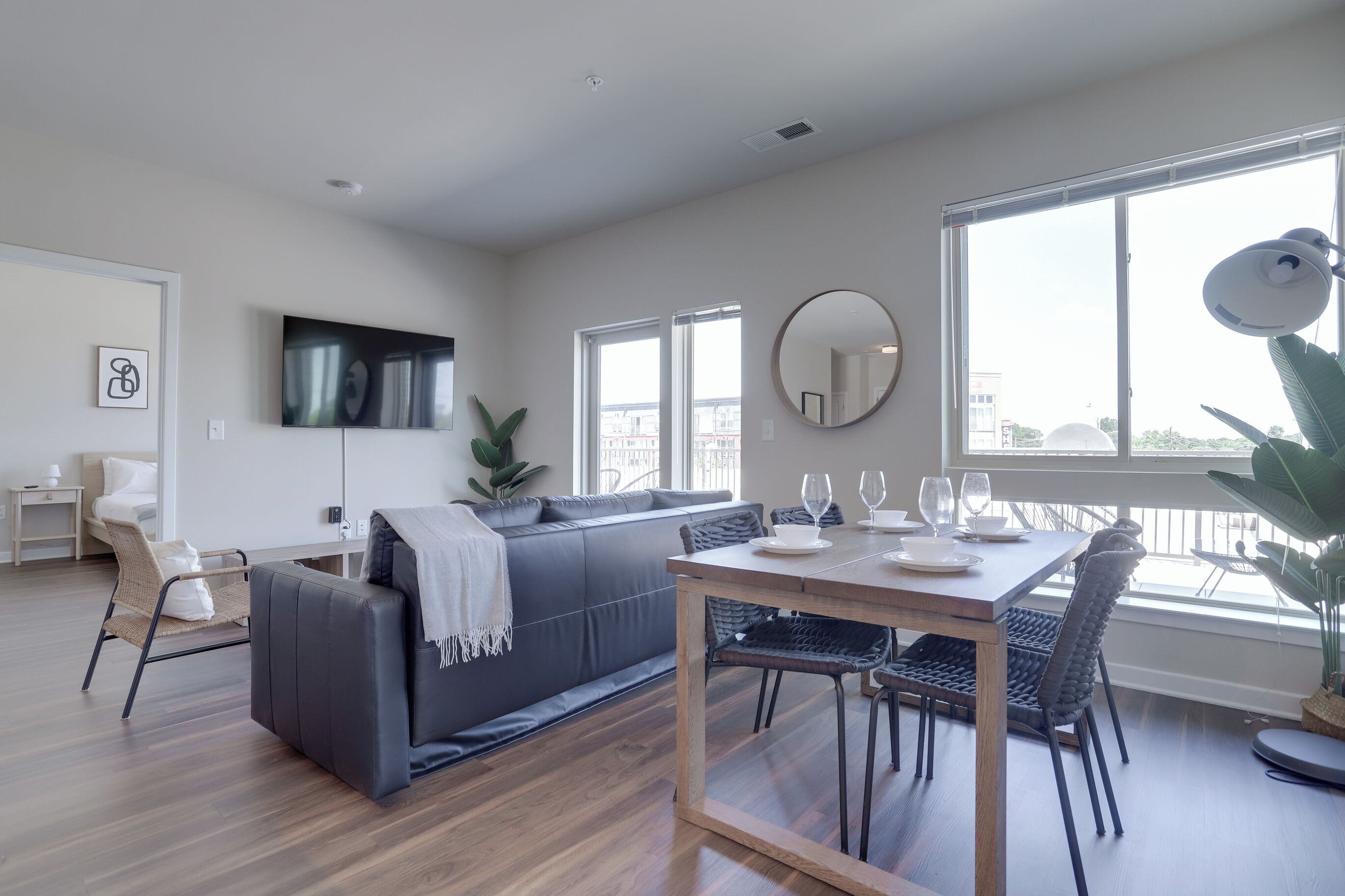 A modern living room with a grey sofa, a dining table with chairs, a large window, and a flat-screen TV mounted on the wall.
