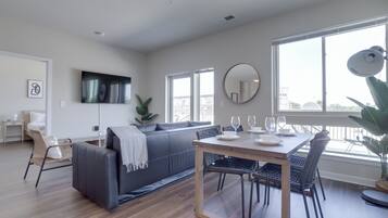 A modern living room with a grey sofa, a dining table with chairs, a large window, and a flat-screen TV mounted on the wall.
