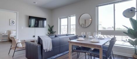 A modern living room with a grey sofa, a dining table with chairs, a large window, and a flat-screen TV mounted on the wall.