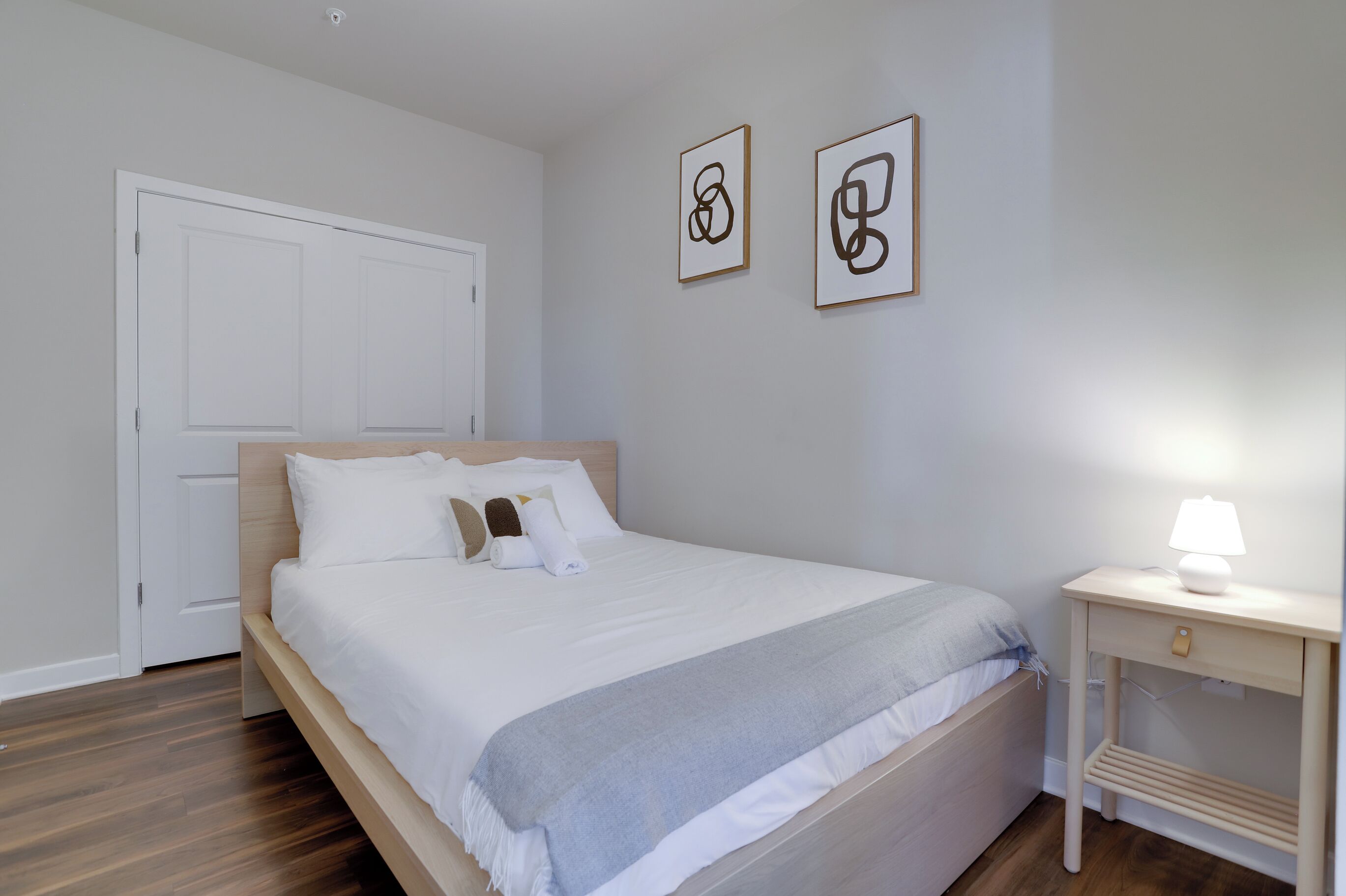 Een net opgemaakt bed met witte beddengoed en een grijze deken, een nachtkastje met een lamp, en twee abstracte kunstwerken aan de muur.