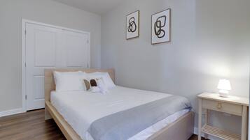 Een net opgemaakt bed met witte beddengoed en een grijze deken, een nachtkastje met een lamp, en twee abstracte kunstwerken aan de muur.