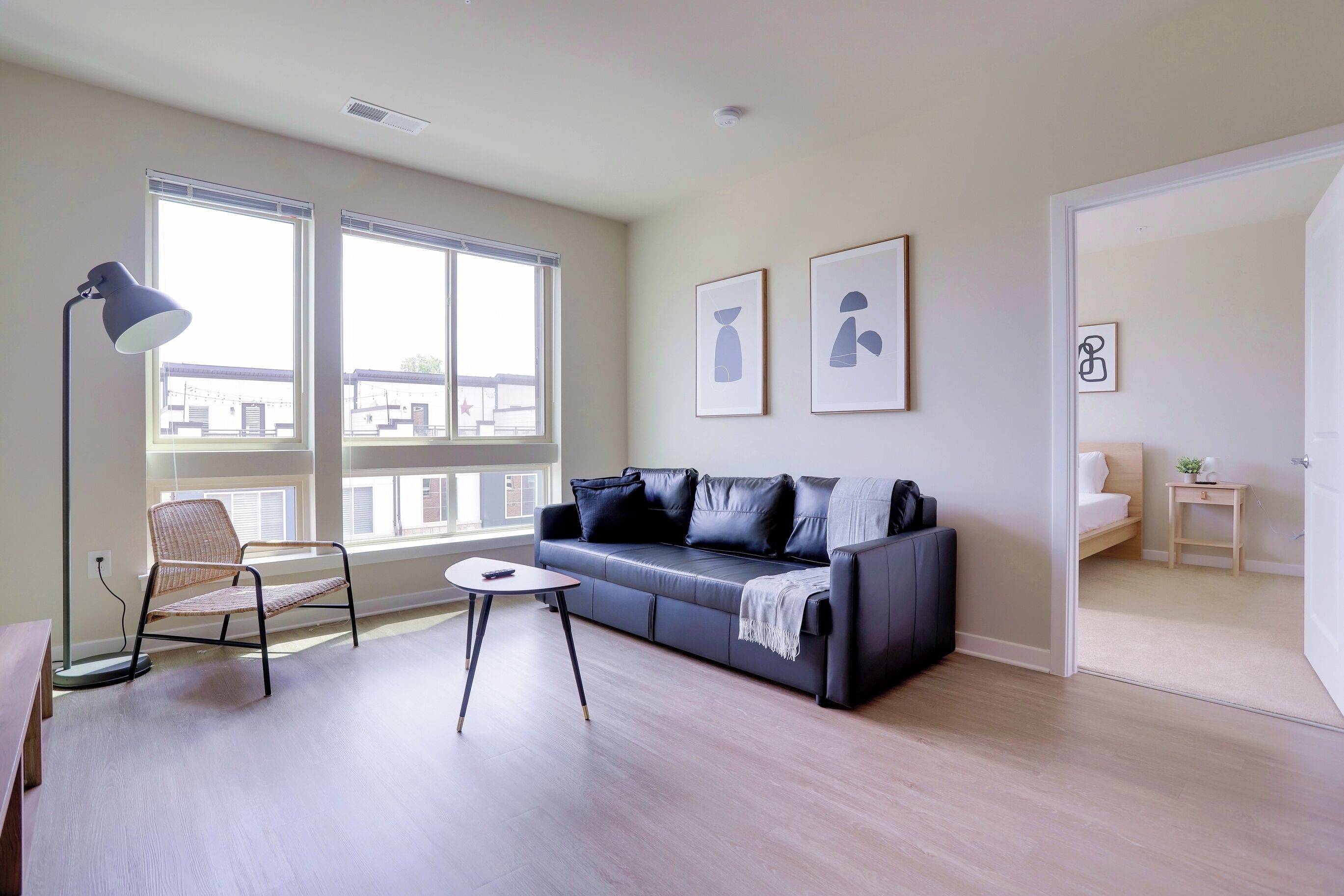 Una sala de estar moderna con un sofá, una butaca, una mesa y una lámpara de pie. Hay ventanas amplias y cuadros en la pared.