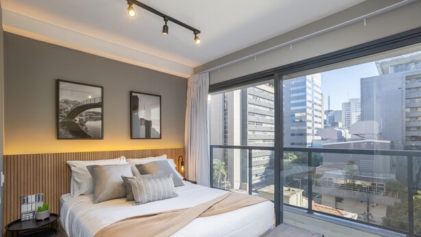 Une chambre d’hôtel moderne avec un grand lit, une petite table de chevet, un banc et une vue sur la ville grâce à de grandes fenêtres.
