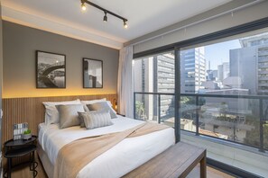 A modern hotel room with a large bed, a small side table, a bench, and a city view through large windows. - Step Up Paraíso (São Paulo)