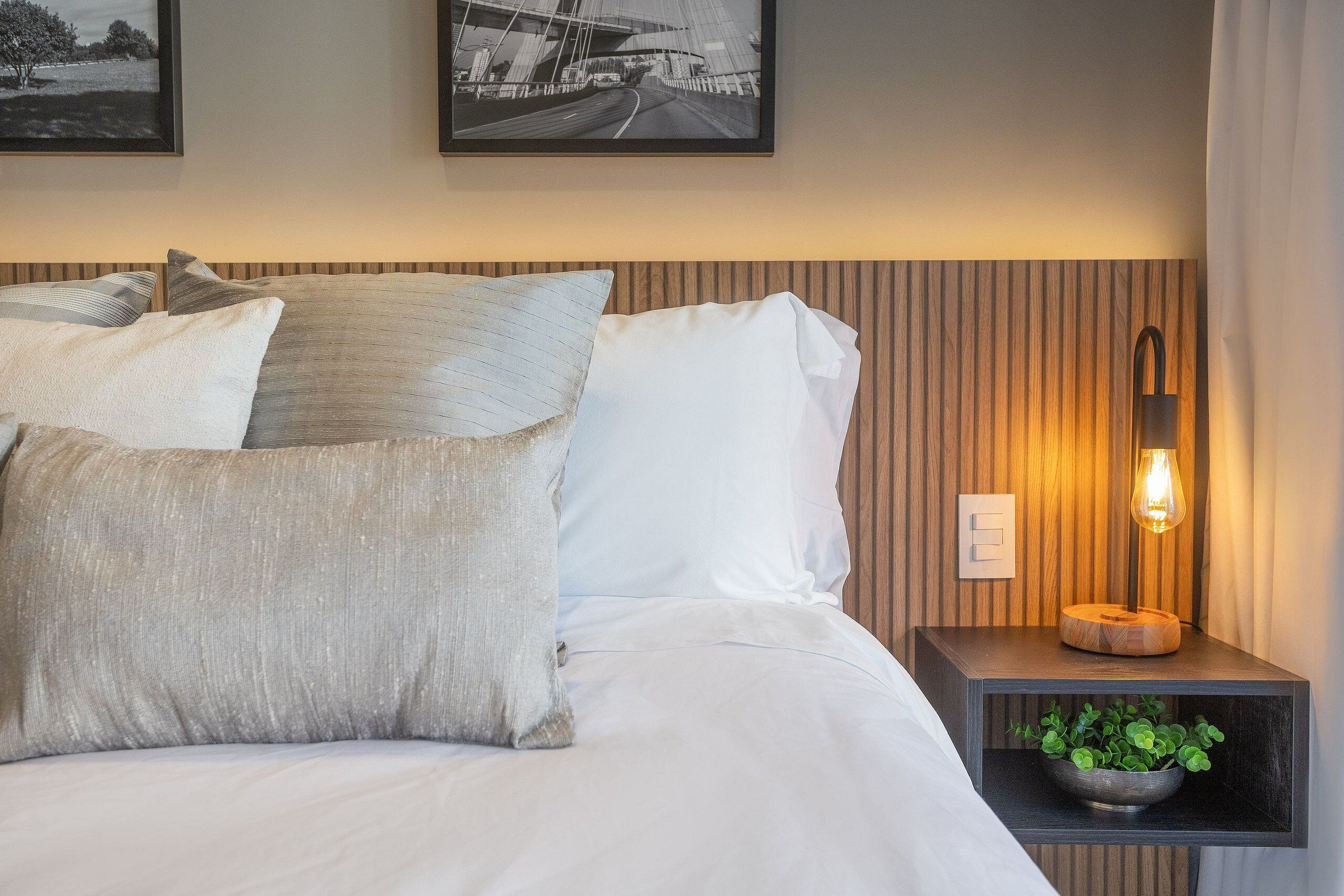 Un lit bien fait, avec du linge de lit blanc et plusieurs oreillers, une table de chevet avec une lampe, et deux tableaux encadrés accrochés au mur.