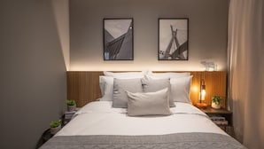 Un lit bien fait, avec des oreillers, une table de chevet surmontée d’une lampe, et deux tableaux encadrés accrochés au mur.