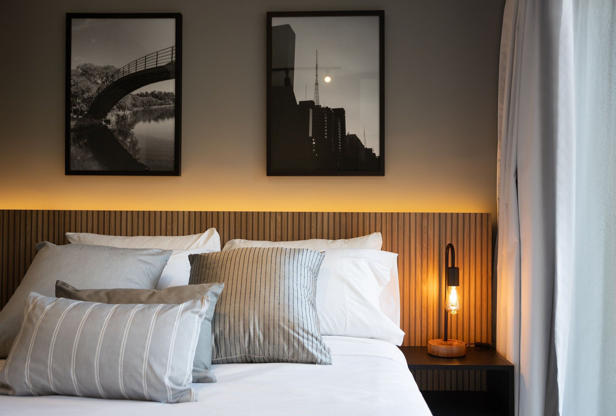 A hotel room with a bed, pillows, a nightstand with a lamp, and two framed pictures on the wall.