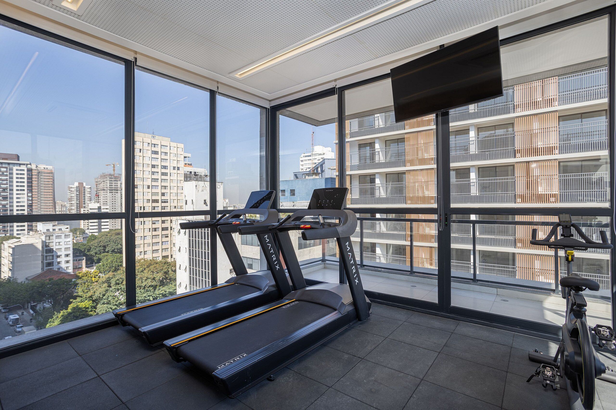 Ein Fitnessstudio mit Laufbändern und einem Ergometer, große Fenster mit Blick auf die Stadt und eine Decke mit einer Leuchte.