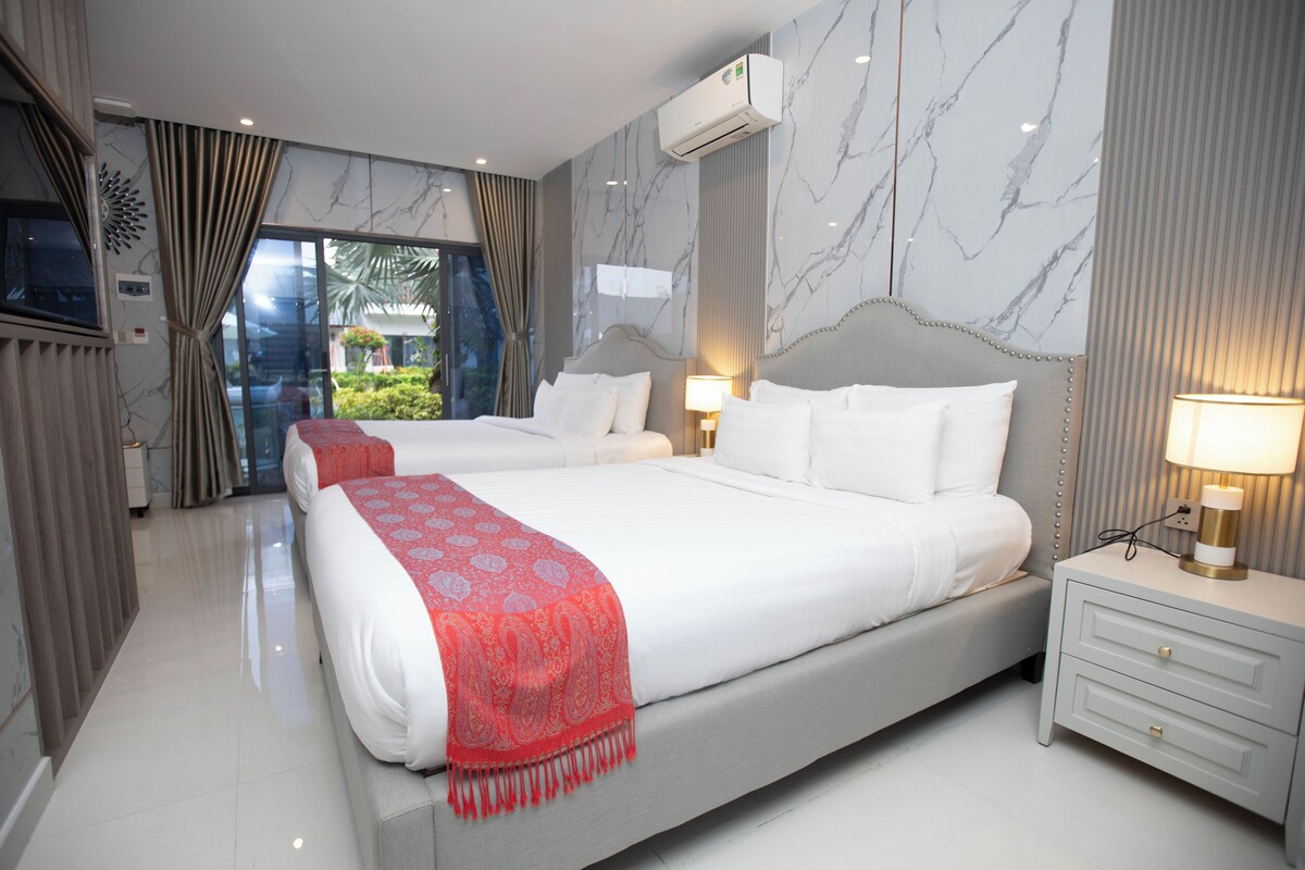 A hotel room with a large bed, a bedside table with a lamp, and a view of greenery through a window.
