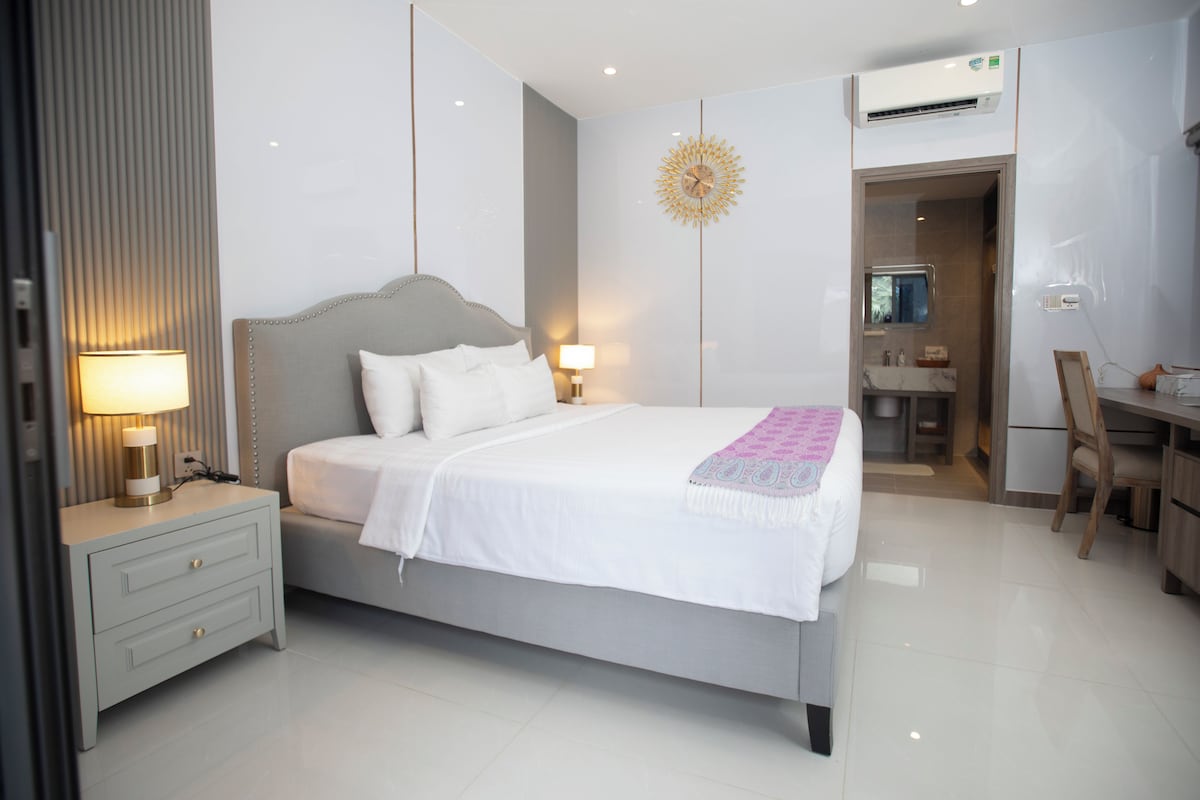 A modern bedroom with a large bed, bedside lamps, a desk, and an air conditioning unit.