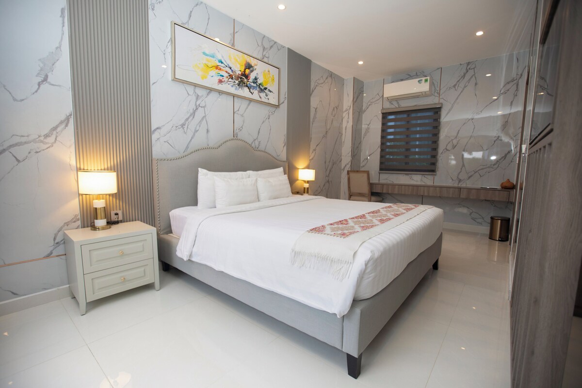 A modern bedroom with a large bed, two bedside tables with lamps, a marble-patterned wall, and a ceiling light.