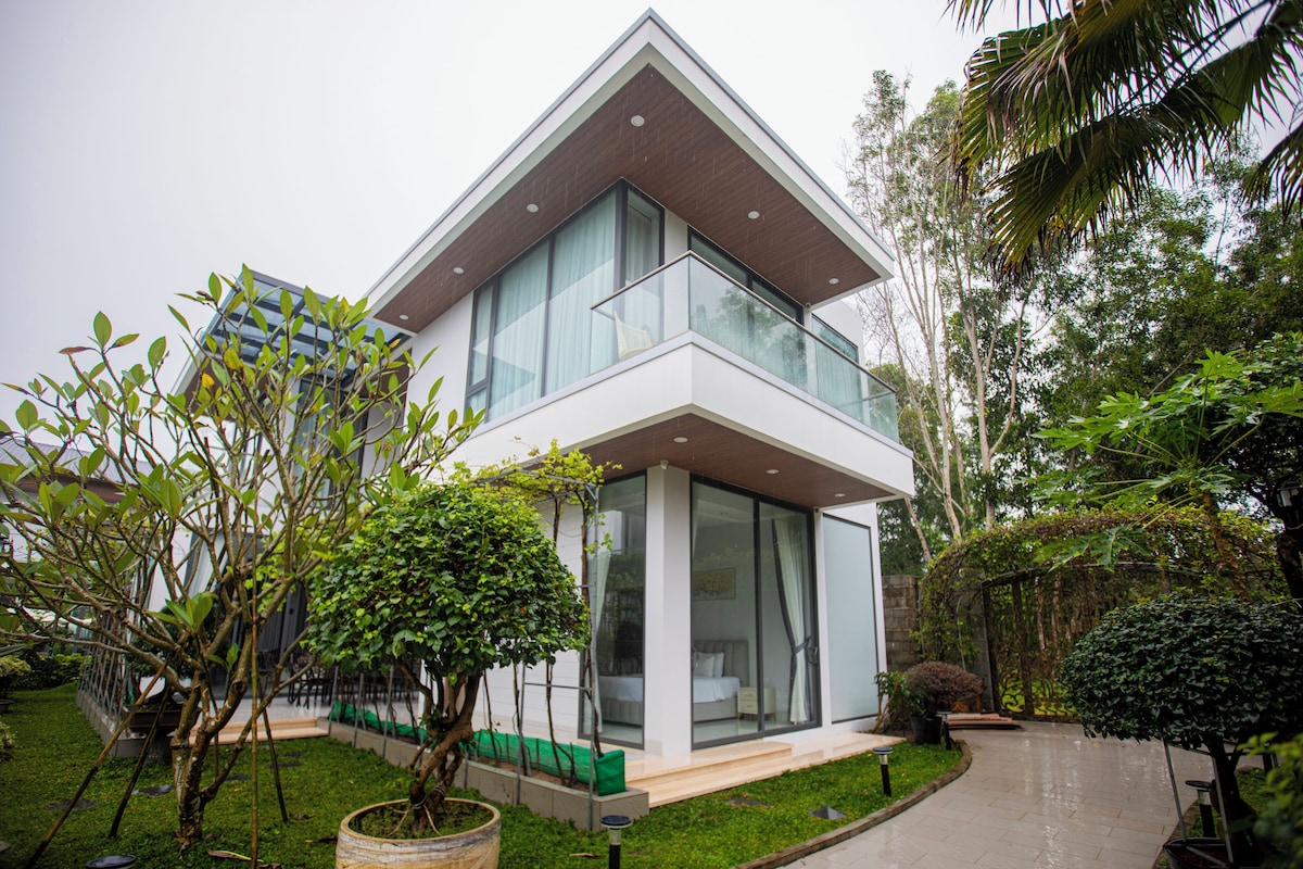A modern two-story house with large glass windows, a well-maintained garden, and a paved pathway.