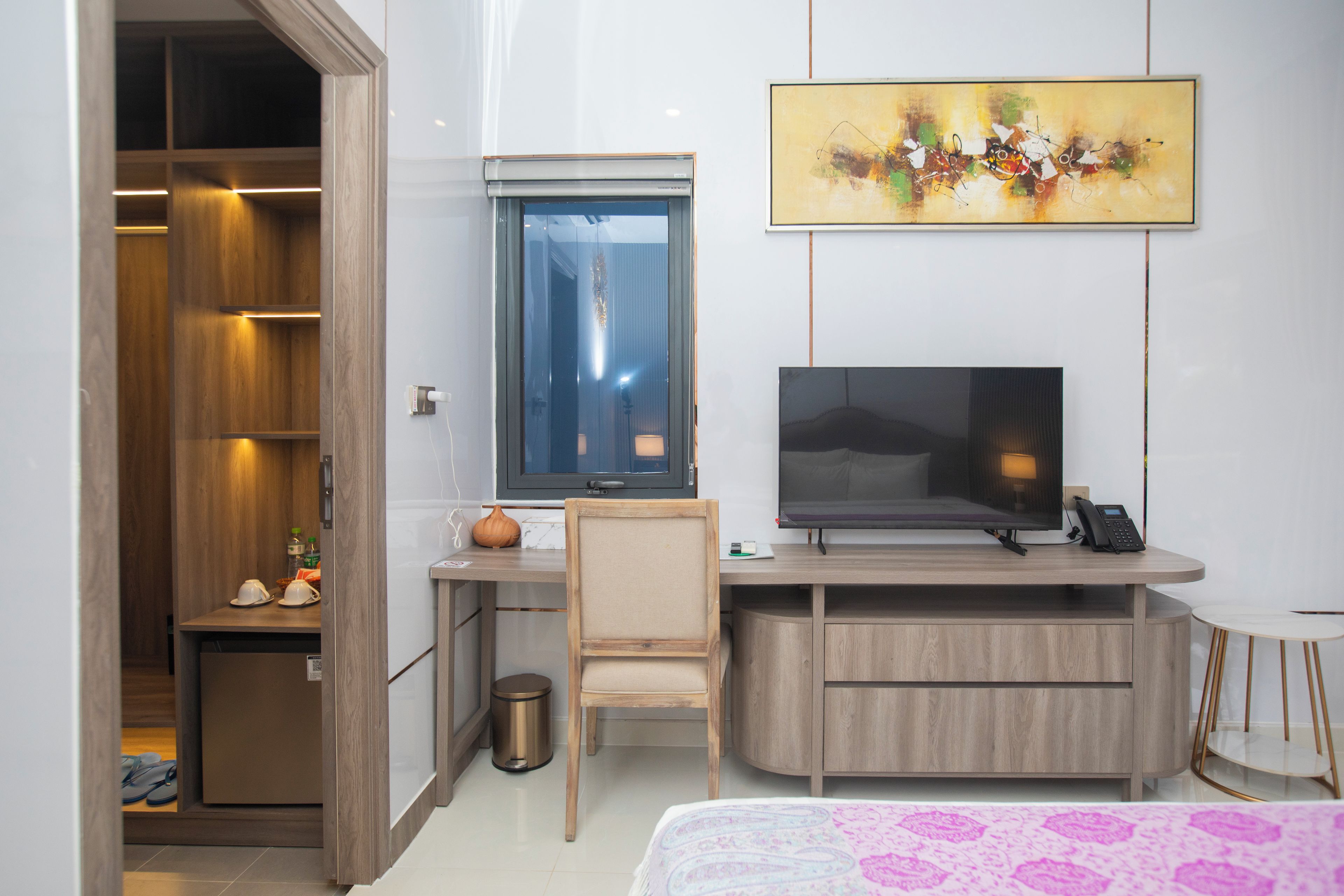 A hotel room with a wooden desk, a television, a small chair, a bedside table, and a bed with a patterned bedspread.