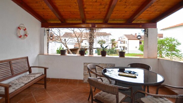 Eine überdachte Terrasse mit Holzboden, einem runden Tisch mit Stühlen, einer Bank und Topfpflanzen.