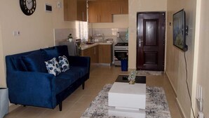 Una sala de estar con un sofá azul, una mesa de centro blanca y una cocina al fondo.