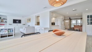 A spacious living area with a large wooden table, white sofas, and a television mounted on the wall.
