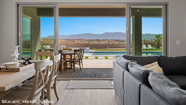 A modern living room with a gray sofa, wooden dining table, and chairs. A glass door leads to an outdoor area with a pool and mountains in the background. - Mountains and Saltwater Pool (Mesquite)