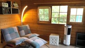 A cozy cabin room with a sofa, a heater, a bookshelf, and a window with a view of greenery.