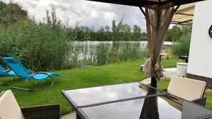 An outdoor patio with a glass table and wicker chairs, overlooking a lake and grassy area.