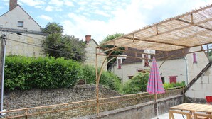 A covered outdoor seating area with a table and chairs, a stone wall, and a building with red shutters in the background.