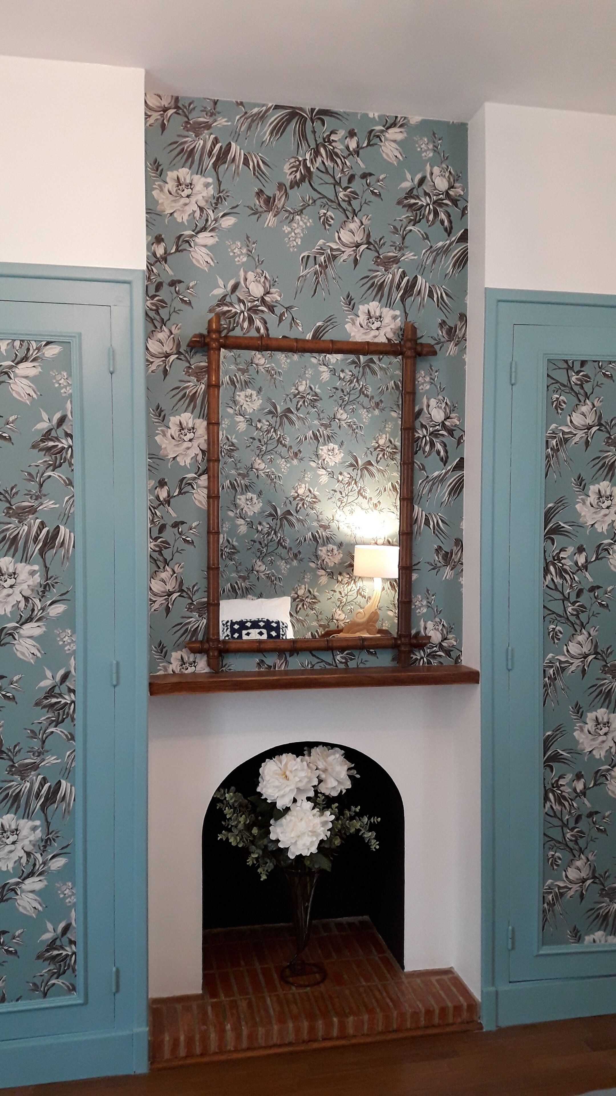 A room with floral wallpaper, a wooden mantel with a mirror, a lit lamp, and a vase with flowers.