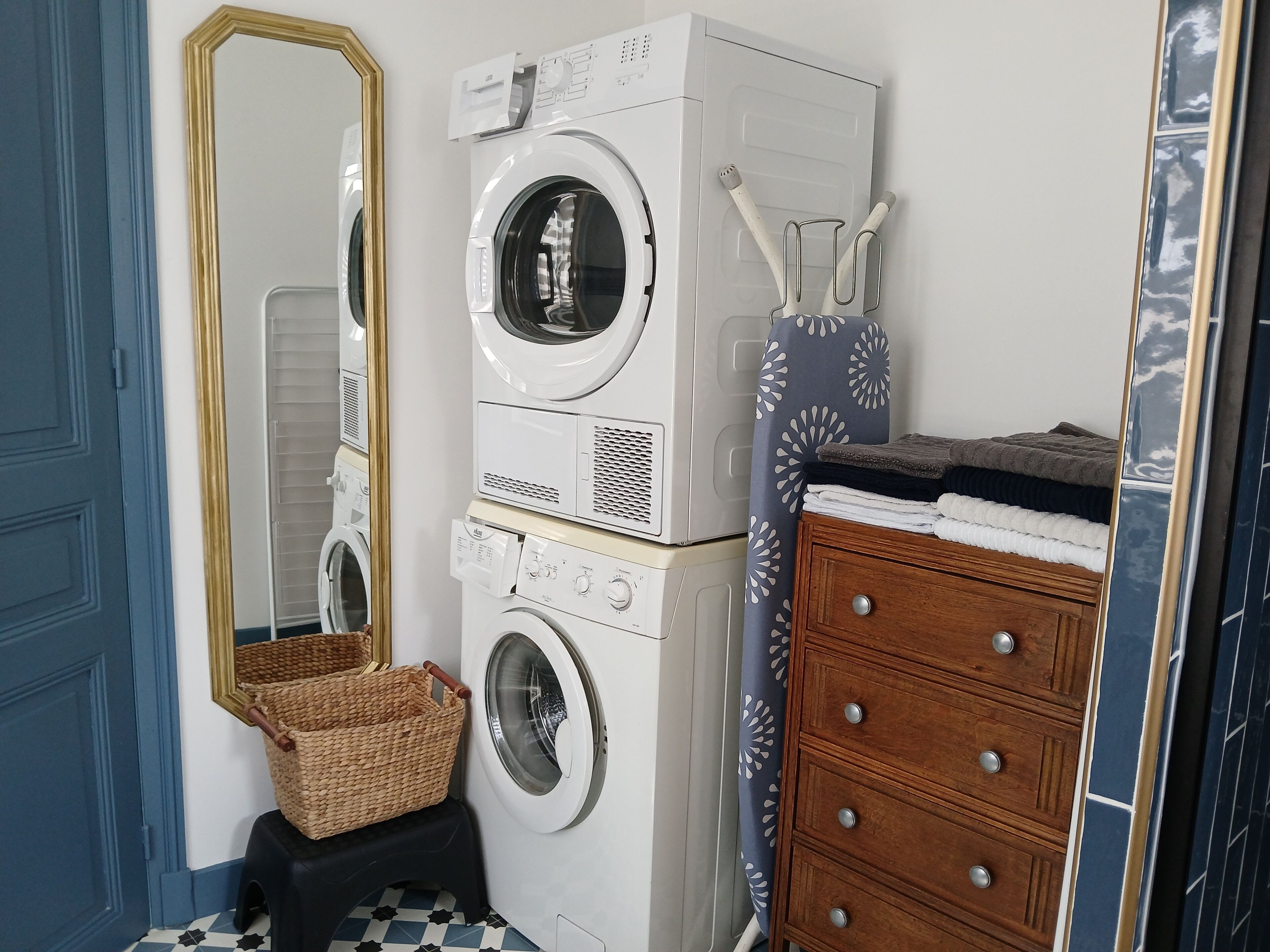 A laundry area with stacked washing machines, a mirror, a wooden dresser, and a basket.