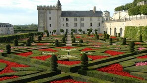 A formal garden with geometrically arranged hedges and flower beds, a large castle-like building, and a clear sky.