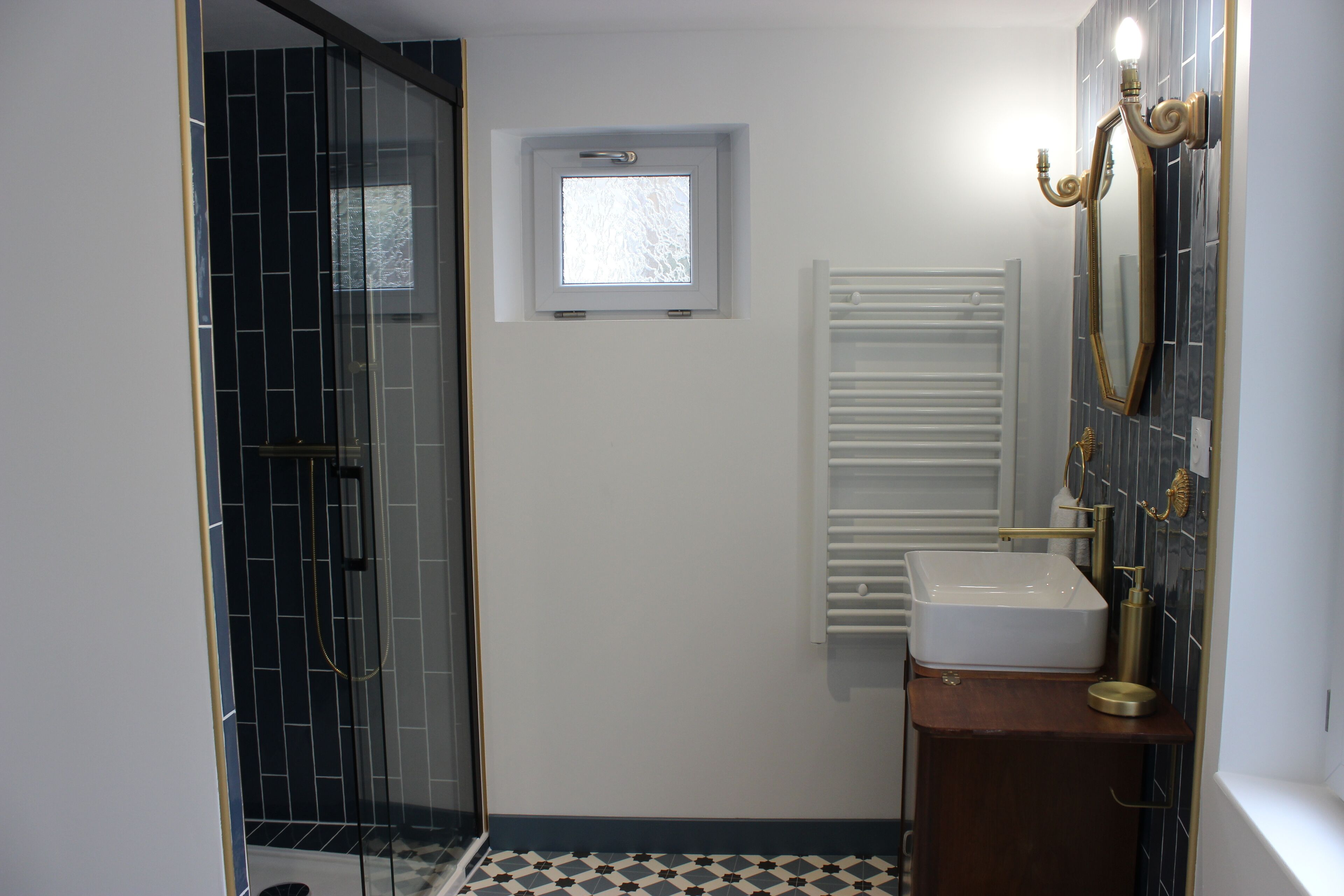Ein Badezimmer mit Duschkabine aus Glas, einem kleinen Fenster, einem weißen Waschbecken und einem Spiegel mit goldener Rahmung.