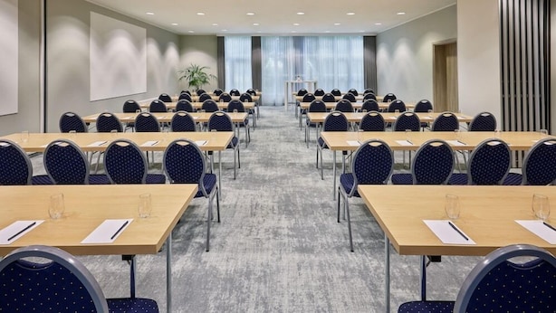 Un salón de conferencias con filas de mesas y sillas, una pantalla grande y una planta.