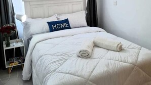 En pænt redt seng med en pude med påskriften 'HOME', i et rum med en hvid sengegavl, et lille natbord med en vase blomster og et billede på væggen.