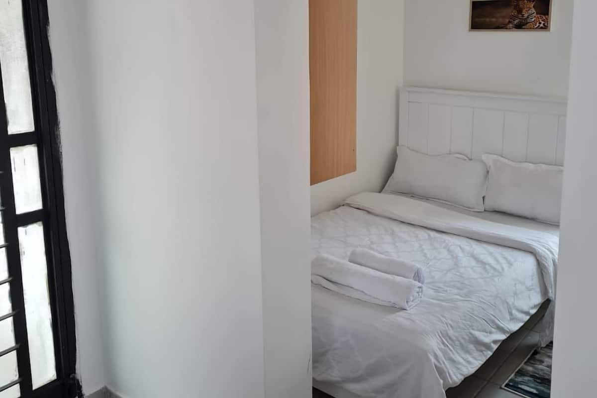 A bedroom with a bed, white walls, a wooden panel, a ceiling light, a framed picture, and a door with a black frame.